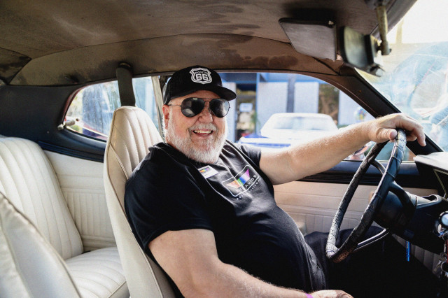 View from the passenger seat of a man sitting in the driver's seat, smiling at the camera, wearing a black hat, sunglasses and black shirt, with one hand on the steering wheel.