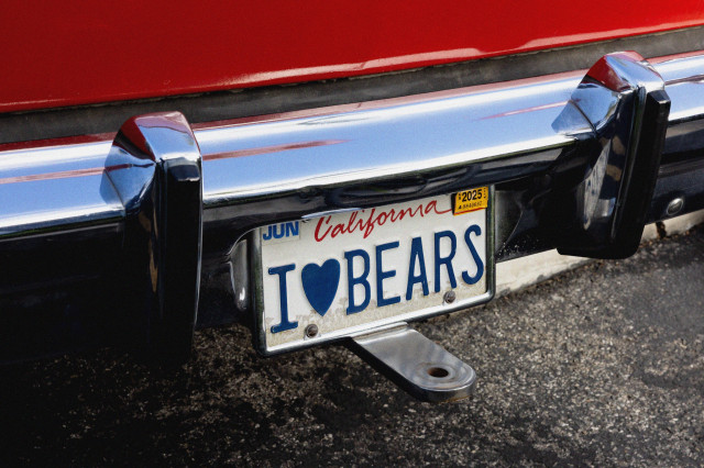 California license plate that reads "I 'heart' BEARS" below a chrome bumper on the back end of a red car.