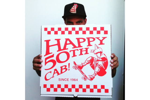 Man in a black baseball cap with a slide of pizza design on it looks over the top of a pizza box (only his eyes are visible) that reads "Happy 50th Cab" in red letters with a design of a skateboarder on the box.