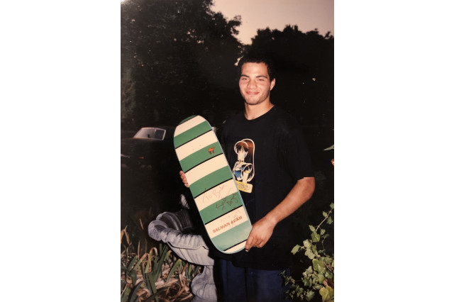 Teenager in a black t-shirt and dark pants, stands in a tree-filled yard, in front of a small, round, cement water fountain, holding a green and white striped skateboard deck.
