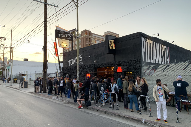 A line of customers wait outside a black brick restaurant at sunset with the name "Pizzanista!" in big white letters on the side of the building.