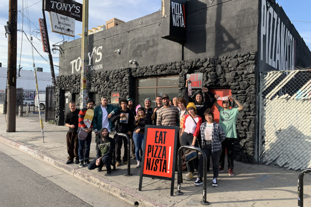 Seventeen young men and women standing behind a red sandwich board sign that says "Eat Pizzanista!", in front of a black brick restaurant with the Pizzanista! logo painted in large white letters on the side. 