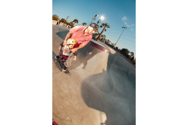 Father in a baseball hat, red shirt and khaki pants on a black skateboard, in a skate park, holding onto the shoulders of his young son who is also on the skateboard, in a yellow helmet, riding between his father's legs.