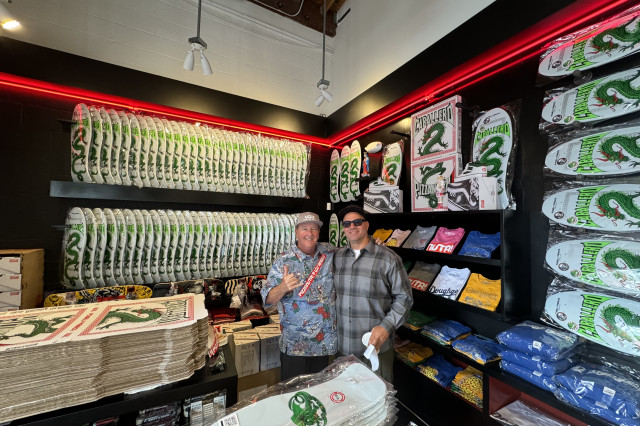 Two men side hugging in a skateboard shop, surrounded by new "Caballero"-branded pizza boxes and skateboard decks, designed with an illustration of a green snake-like dragon. 