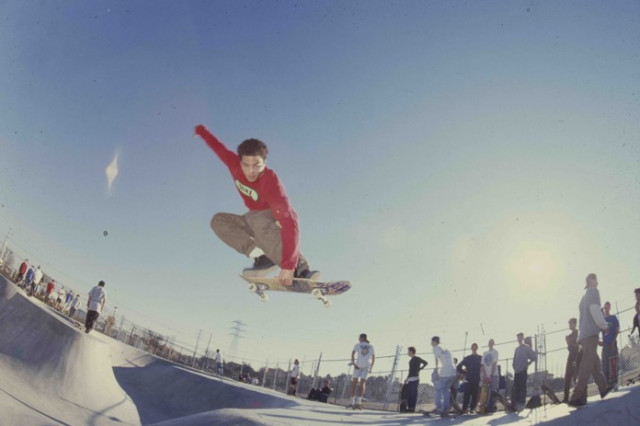 Young man in long-sleeved red shirt and khaki pants with one hand up in teh air and the other crabbing his skateboard as he jumps in midair above a skate ramp.