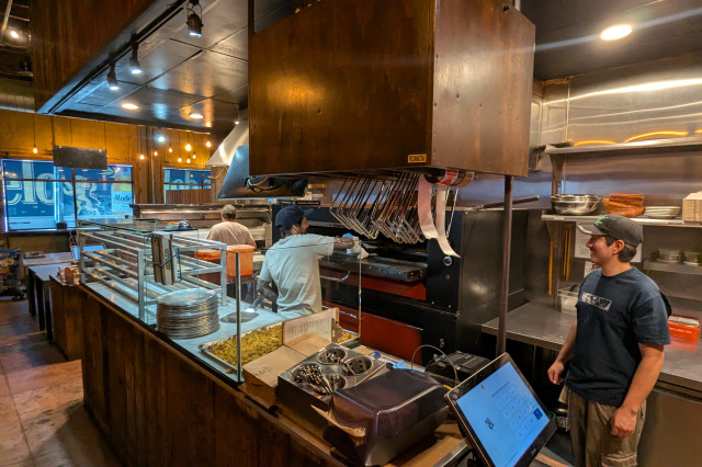 Inside a small restaurant with an open kitchen, two men in white shorts prepping pizzas and sliding them into a large clay oven, while a third man in a black shirt stands off to the side chatting with his busy coworkers.