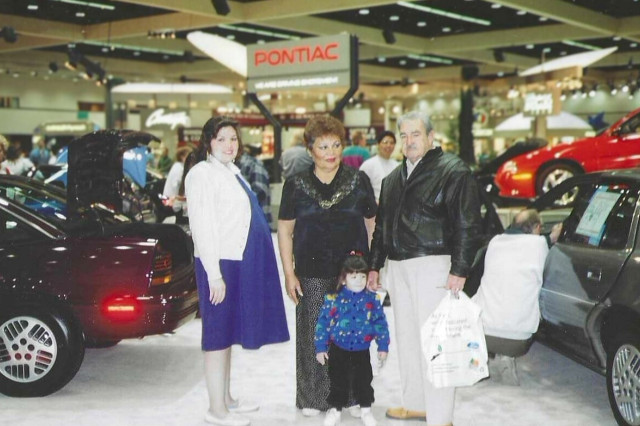 A pregnant woman, an older couple and a young girl stand at a car show.