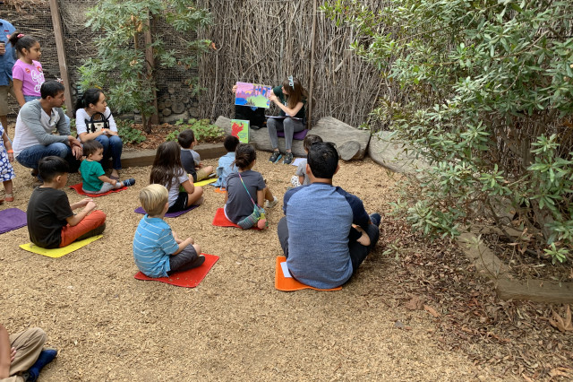 A group of children and adults sitting on individual mats on a wood chip ground, listening to a person read a large picture book during an outdoor story time session. 