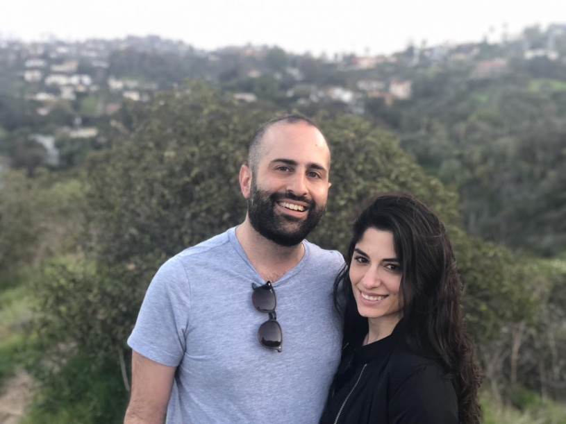 A smiling couple facing the camera stand in nature with a view of the city in the background